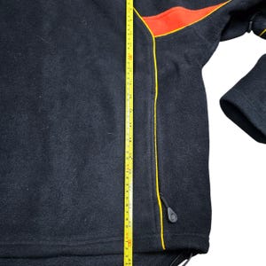 May include: Black fleece jacket with red and yellow detailing. The jacket features a zip and a drawstring. A measuring tape indicates the jacket is approximately 61 cm long.