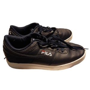 May include: A pair of navy blue Fila sneakers with white soles and laces. The Fila logo is displayed in white with a red accent. The shoes have a low-profile design and are made of a textured material.
