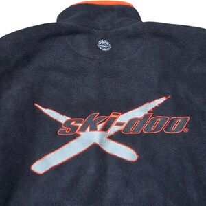 May include: Black fleece jacket with a high collar and orange trim. The front features the word "ski-doo" in silver with an orange outline, and a stylized "X" design. A small logo is visible on the upper back.