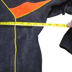 May include: Black fleece jacket with orange and yellow details. A tape measure shows a measurement of approximately 63.5 cm. The jacket is being measured.