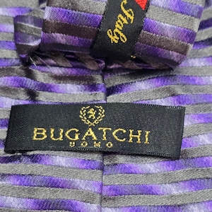 May include: A necktie featuring a pattern of alternating purple and dark gray horizontal stripes. A black fabric label is stitched onto the tie, displaying the gold text BUGATCHI UOMO with a small crest icon above the brand name and Italy text.