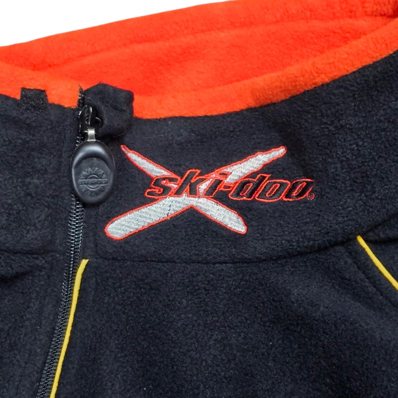 May include: Close-up of a black fleece jacket with an orange collar. The jacket features a zipper and the "Ski-doo" logo embroidered in red and silver. Yellow stitching accents the design.