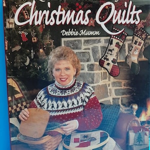 Könnte beinhalten: Buchcover von "Quick Country Christmas Quilts" von Debbie Mumm. Es zeigt eine Frau in einem rot-weiß-blauen Pullover, die einen Korb und einen Stickrahmen hält. Ein Weihnachtsbaum und Strümpfe sind im Hintergrund zu sehen.