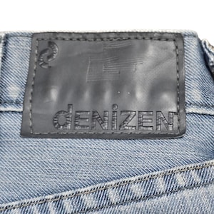 Puede incluir: Primer plano de una etiqueta de vaqueros de mezclilla. La etiqueta es negra con la palabra "DENIZEN" en relieve en una fuente sans-serif. La etiqueta est&aacute; cosida a la tela vaquera azul.