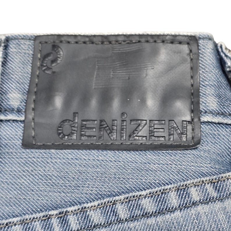 Puede incluir: Primer plano de una etiqueta de vaqueros de mezclilla. La etiqueta es negra con la palabra "DENIZEN" en relieve en una fuente sans-serif. La etiqueta est&aacute; cosida a la tela vaquera azul.