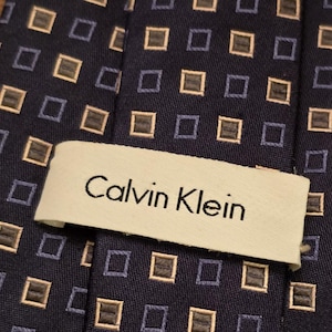 Puede incluir: Una corbata azul oscuro con un patrón repetitivo de pequeños cuadrados en beige y azul claro. La corbata tiene una etiqueta blanca con el texto "Calvin Klein" en negro.