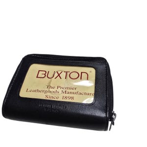 May include: Black leather wallet with a zipper closure. The wallet features a clear window with the brand name "BUXTON" and the text "The Premier Leathergoods Manufacture Since 1898".