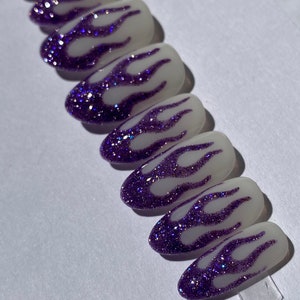 Puede incluir: Un conjunto de diez u&ntilde;as postizas ovaladas con base blanca y dise&ntilde;o de llamas de purpurina morada.