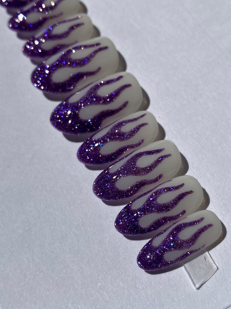Puede incluir: Un conjunto de diez u&ntilde;as postizas ovaladas con base blanca y dise&ntilde;o de llamas de purpurina morada.