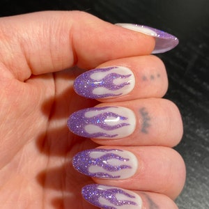 Puede incluir: Primer plano de una mano con cinco u&ntilde;as ovaladas pintadas con una base blanca y un dise&ntilde;o de llama de purpurina morada.