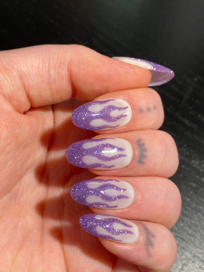 Puede incluir: Primer plano de una mano con cinco u&ntilde;as ovaladas pintadas con una base blanca y un dise&ntilde;o de llama de purpurina morada.