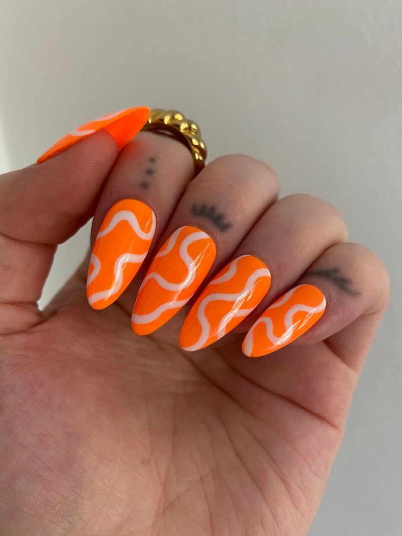 May include: Orange and white swirl patterned press-on nails. The nails are long and have a pointed shape.