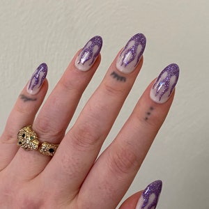 Puede incluir: Primer plano de una mano con u&ntilde;as largas y ovaladas pintadas con una base blanca y un dise&ntilde;o de llama de purpurina morada. La mano lleva un anillo de oro con una piedra negra y verde.