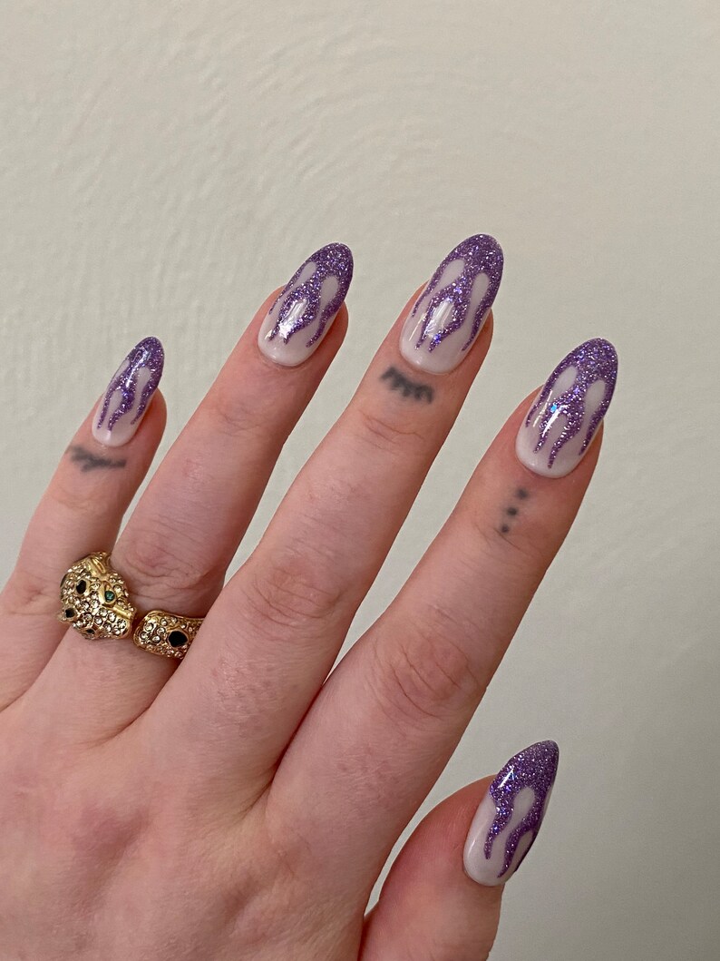 Puede incluir: Primer plano de una mano con u&ntilde;as largas y ovaladas pintadas con una base blanca y un dise&ntilde;o de llama de purpurina morada. La mano lleva un anillo de oro con una piedra negra y verde.