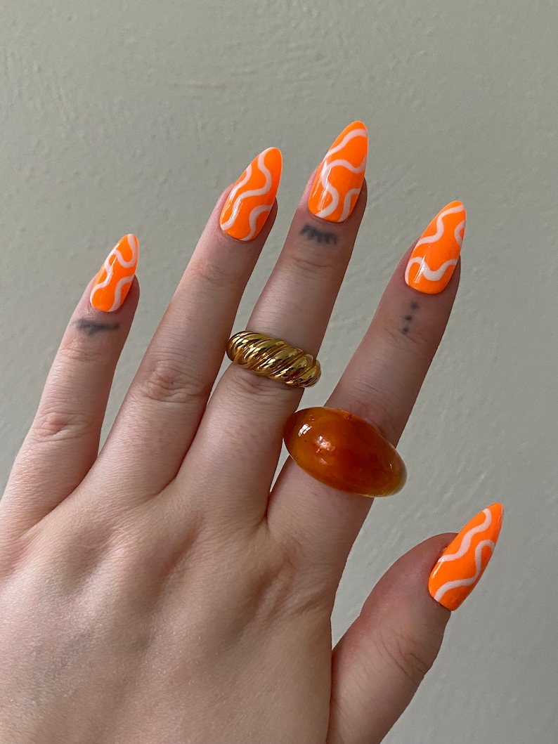 May include: Orange almond shaped press-on nails with a white wavy design. The nails are shown on a hand wearing two rings, one gold and one amber.