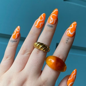 May include: Close-up of a hand with orange and white swirl nail polish. The hand is wearing two rings, one gold and one amber.
