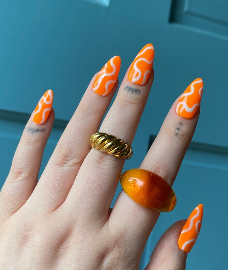 May include: Close-up of a hand with orange and white swirl nail polish. The hand is wearing two rings, one gold and one amber.