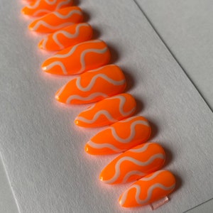 May include: Set of ten orange and white false nails with a wavy design. The nails are long and have a pointed shape.