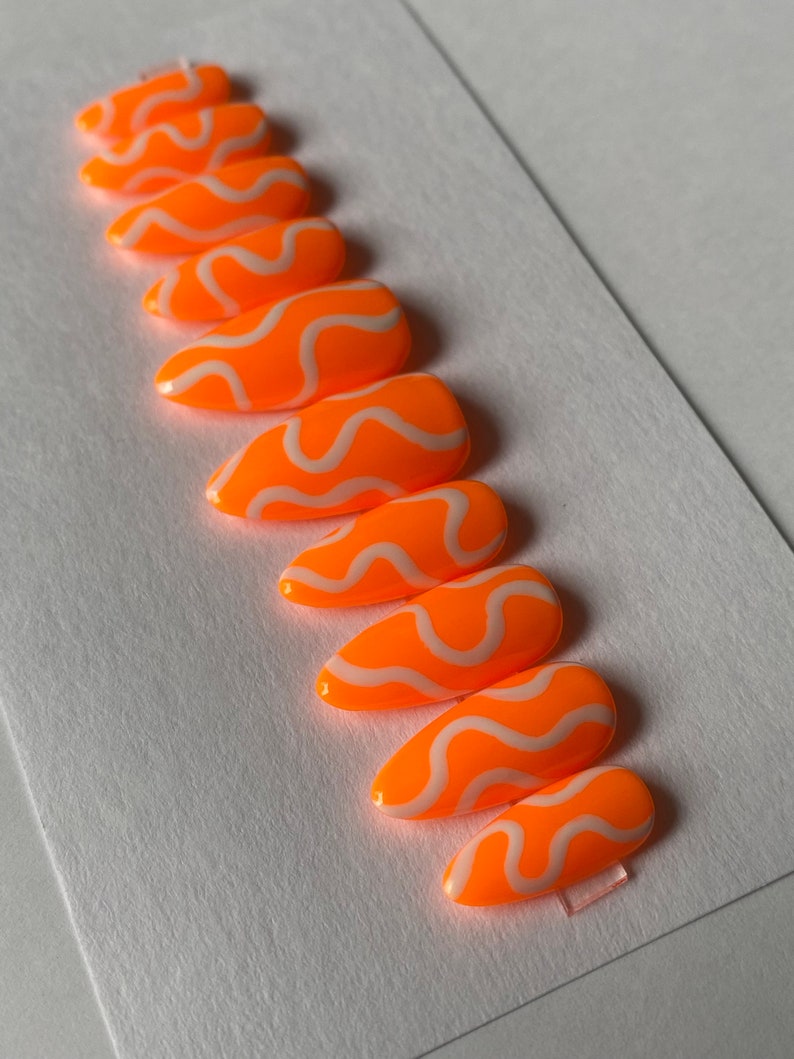 May include: Set of ten orange and white false nails with a wavy design. The nails are long and have a pointed shape.