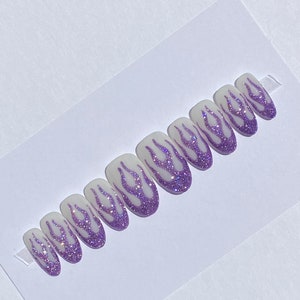 Puede incluir: Conjunto de diez u&ntilde;as postizas con base blanca y dise&ntilde;o de llamas de purpurina morada.