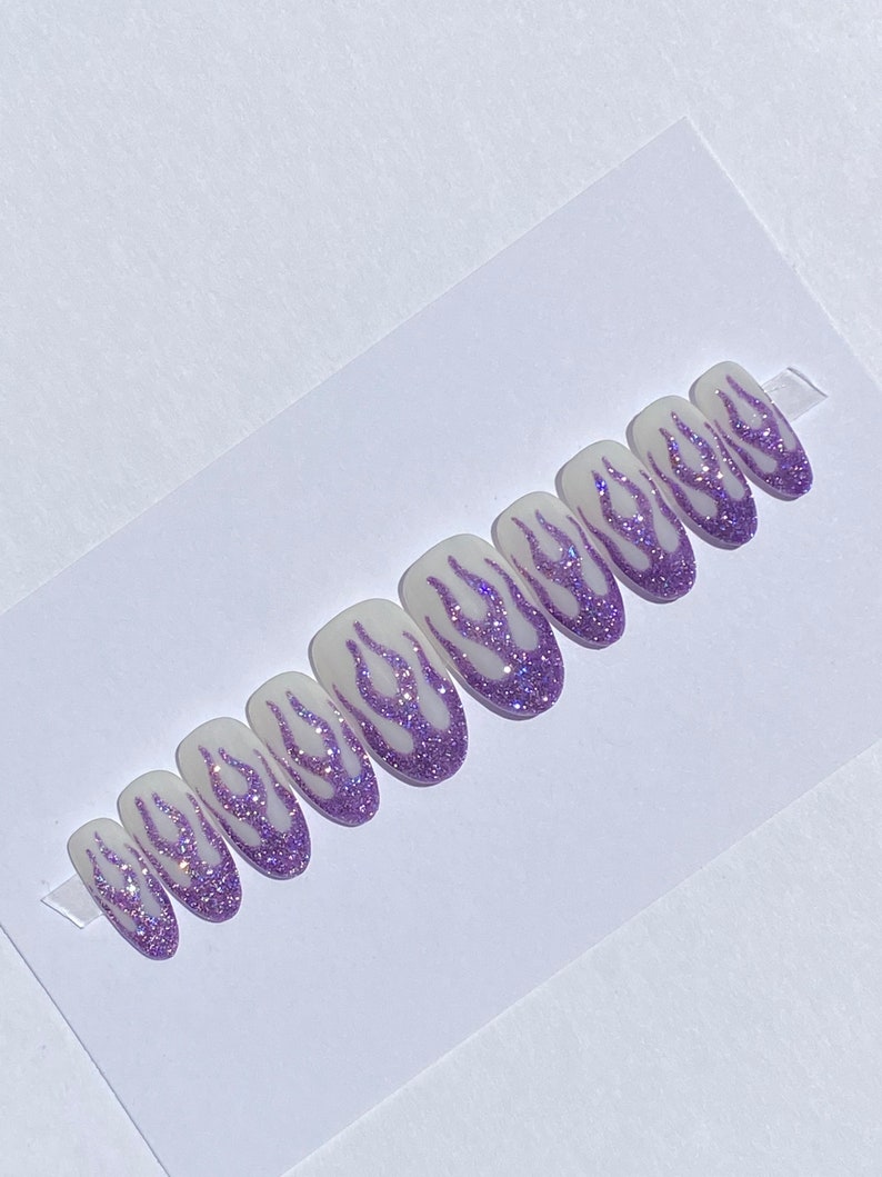 Puede incluir: Conjunto de diez u&ntilde;as postizas con base blanca y dise&ntilde;o de llamas de purpurina morada.