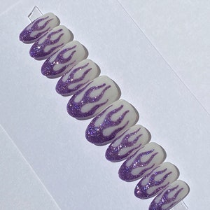 Puede incluir: Conjunto de diez u&ntilde;as postizas con base blanca y dise&ntilde;o de llamas de purpurina morada.