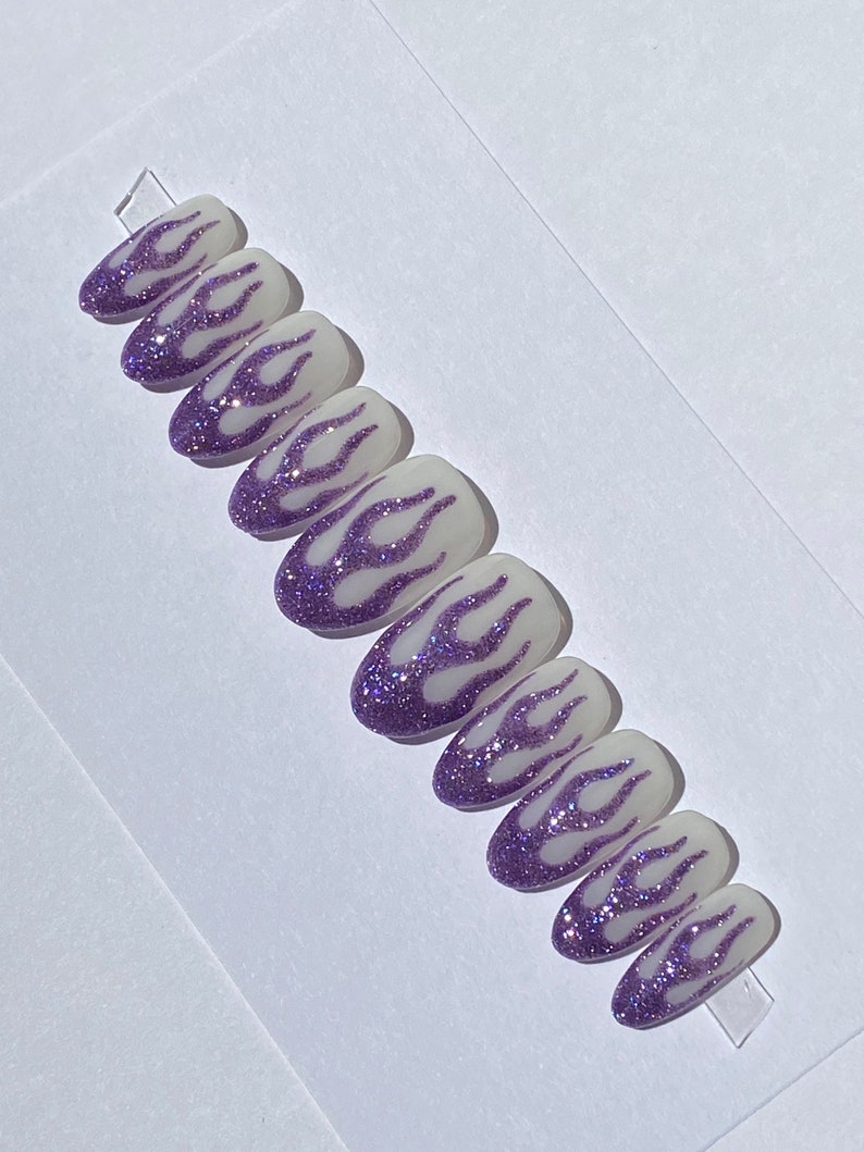 Puede incluir: Conjunto de diez u&ntilde;as postizas con base blanca y dise&ntilde;o de llamas de purpurina morada.