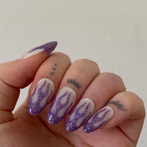 Puede incluir: U&ntilde;as postizas ovaladas blancas con un dise&ntilde;o de llamas de purpurina morada.