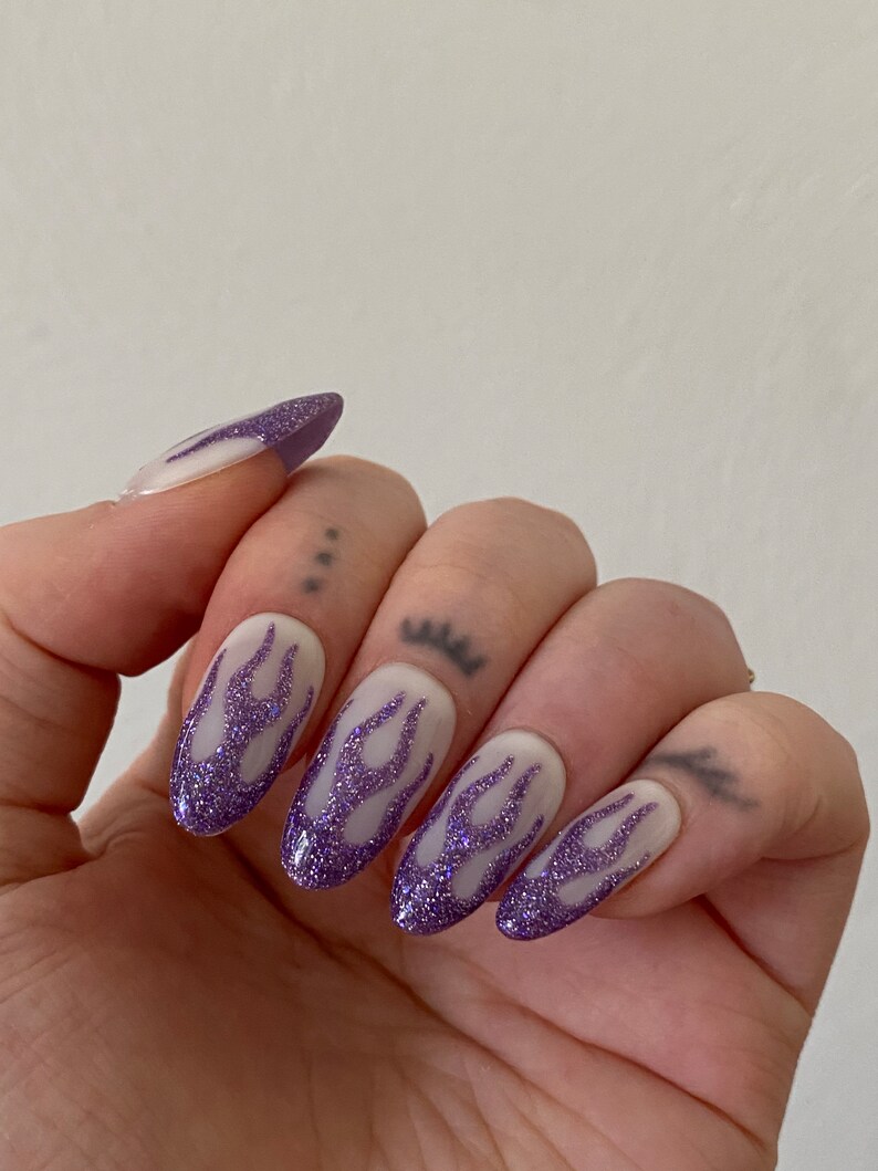 Puede incluir: U&ntilde;as postizas ovaladas blancas con un dise&ntilde;o de llamas de purpurina morada.