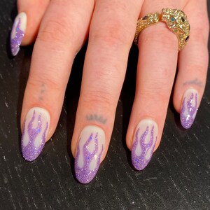 Puede incluir: Primer plano de una mano con cinco u&ntilde;as pintadas de blanco con un dise&ntilde;o de llama de purpurina morada. El dedo anular tiene un anillo de oro con un dise&ntilde;o de pantera enjoyado.