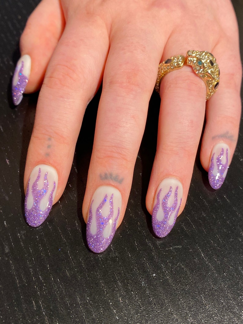 Puede incluir: Primer plano de una mano con cinco u&ntilde;as pintadas de blanco con un dise&ntilde;o de llama de purpurina morada. El dedo anular tiene un anillo de oro con un dise&ntilde;o de pantera enjoyado.