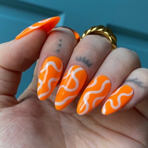 May include: Orange almond-shaped press-on nails with white wavy lines.