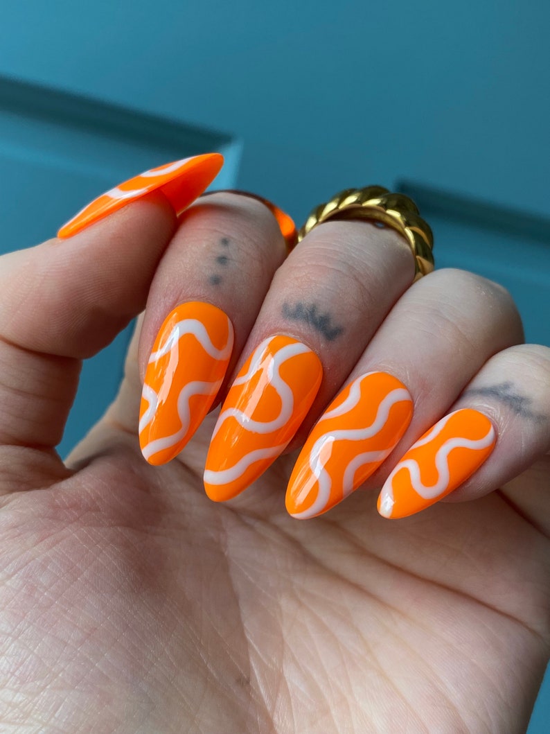 May include: Orange almond-shaped press-on nails with white wavy lines.
