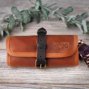 K&ouml;nnte beinhalten: Eine braune Lederuhrentasche mit einem schwarzen Riemen und dem Namen "KLEO" gepr&auml;gt auf der Vorderseite. Eine violette Uhr mit einem Metallband liegt neben der Tasche.