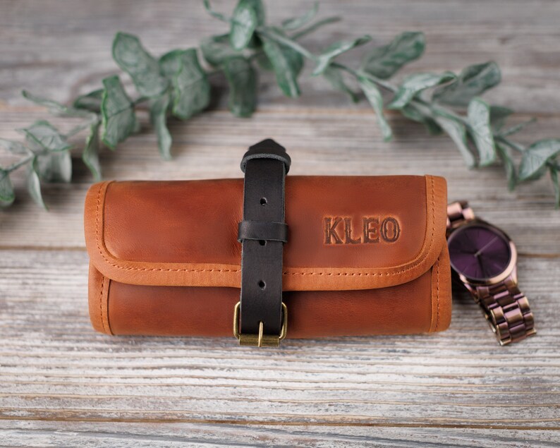 K&ouml;nnte beinhalten: Eine braune Lederuhrentasche mit einem schwarzen Riemen und dem Namen "KLEO" gepr&auml;gt auf der Vorderseite. Eine violette Uhr mit einem Metallband liegt neben der Tasche.