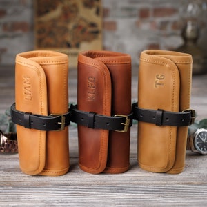 K&ouml;nnte beinhalten: Drei personalisierte Leder-Uhrenrollen in Braun, Tan und Hellbraun mit schwarzen Riemen und den Namen "LLAM", "KLEO" und "TG" auf der Vorderseite gepr&auml;gt.