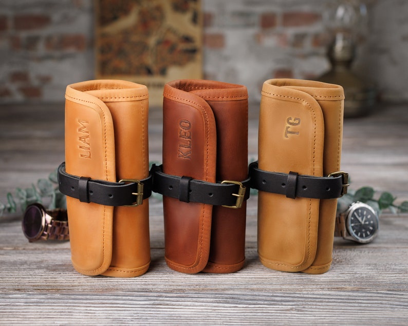 K&ouml;nnte beinhalten: Drei personalisierte Leder-Uhrenrollen in Braun, Tan und Hellbraun mit schwarzen Riemen und den Namen "LLAM", "KLEO" und "TG" auf der Vorderseite gepr&auml;gt.
