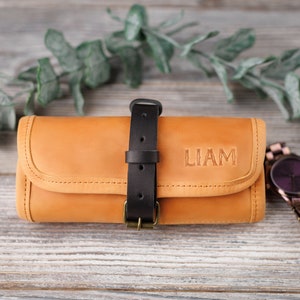 K&ouml;nnte beinhalten: Eine braune Lederuhrrolle mit einem schwarzen Riemen und einer Schnalle. Die Rolle ist mit dem Namen "LIAM" in schwarzen Buchstaben personalisiert.