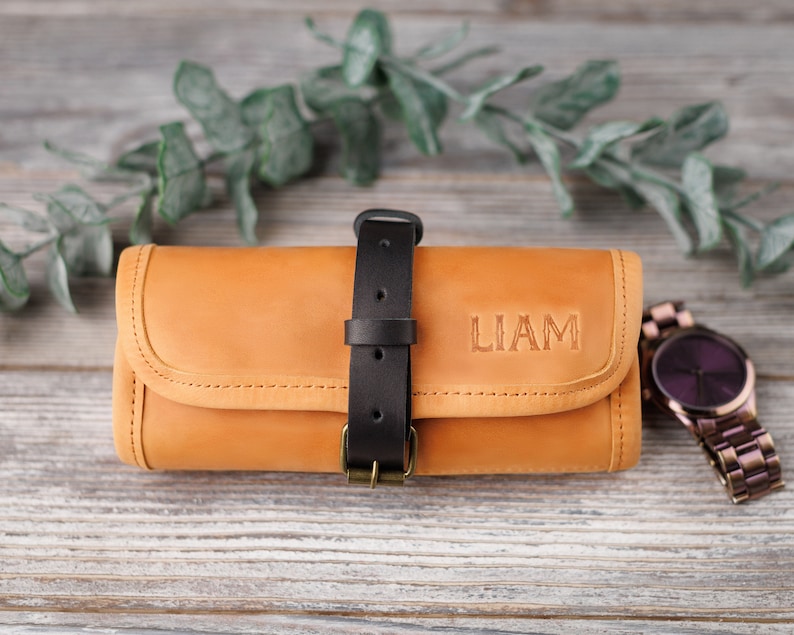 K&ouml;nnte beinhalten: Eine braune Lederuhrrolle mit einem schwarzen Riemen und einer Schnalle. Die Rolle ist mit dem Namen "LIAM" in schwarzen Buchstaben personalisiert.