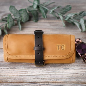 K&ouml;nnte beinhalten: Eine braune Lederuhrrolle mit einem schwarzen Riemen und einer Schnalle. Die Rolle hat ein Monogramm "TG" auf der Vorderseite.