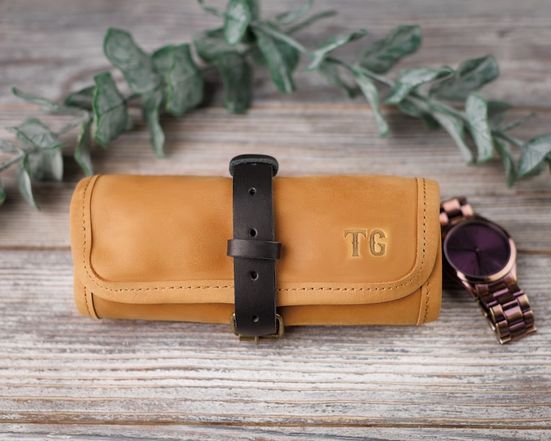 K&ouml;nnte beinhalten: Eine braune Lederuhrrolle mit einem schwarzen Riemen und einer Schnalle. Die Rolle hat ein Monogramm "TG" auf der Vorderseite.