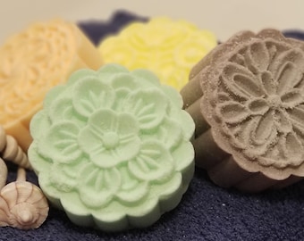 May include: A collection of decorative soaps in various colors, including yellow, green, and brown, with floral designs. The soaps are arranged on a dark blue towel, accompanied by seashells and a porous rock.