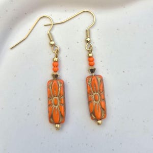 May include: A pair of drop earrings featuring rectangular orange beads with a gold-toned floral pattern. Each earring includes a small orange round bead, a faceted gold-toned bead, and a gold-toned hook wire for attachment to the ear.