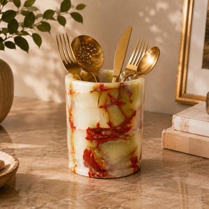 May include: A cylindrical stone utensil holder featuring a polished finish with a pale green base colour and prominent, irregular deep red veining. The container is filled with a set of gold-toned metal cutlery, including forks, a knife, and spoons.