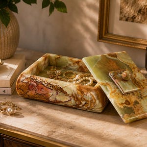 Puede incluir: Un joyero rectangular hecho de piedra ónix pulida en tonos verdes y marrones. La caja cuenta con una tapa extraíble con un asa cuadrada y una superficie lisa y veteada. En su interior, la caja contiene varios anillos y collares dorados.