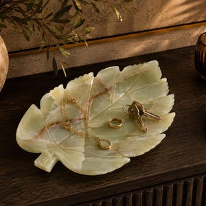 Può includere: Un vassoio in pietra traslucida verde pallido a forma di grande foglia con venature visibili e inclusioni marroni naturali. Il vassoio contiene una collana a catena color oro, un semplice anello d'oro e due chiavi metalliche su un anello.
