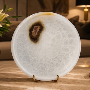 May include: A circular white stone tray with a translucent, marbled appearance and a prominent dark brown, textured inclusion near the top edge. The tray is supported by a small, gold-toned metal stand, showcasing its smooth, polished surface.