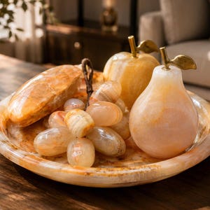 Op de afbeelding: Een decoratieve set van stenen fruit met een gepolijste oranje en crèmekleurige onyx appel, een peer en een tros druiven op een bijpassend rond stenen dienblad. Elk stuk fruit is voorzien van een goudkleurige metalen steel.