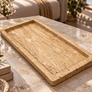 Puede incluir: Una bandeja rectangular hecha de piedra travertino beige, con una textura porosa natural y una superficie plana y lisa con bordes elevados. La bandeja muestra vetas características de color marrón claro y crema en toda su estructura.