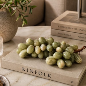 May include: A decorative cluster of grapes carved from polished light green stone, featuring smooth, rounded individual grape shapes with natural veining and a brown wrapped stem, resting on a beige book with the word KINFOLK printed on the cover.